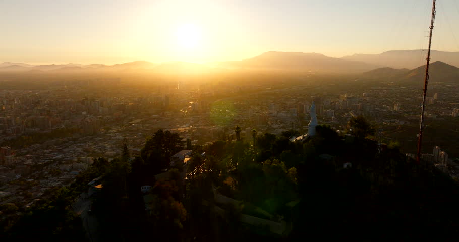 Cerro San Cristobal hill with Virgin Mary statue, overlooking Santiago cityscape at golden hour, Chile. Aerial forward