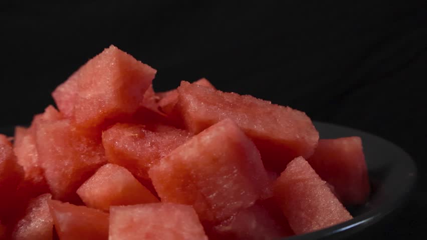 Close up video of fresh and tasty red colored watermelon sliced cubes in a bowl while placed in a pile in front of a black and dark blurry bokeh background. Juicy fruit ready to be eaten and snacked.