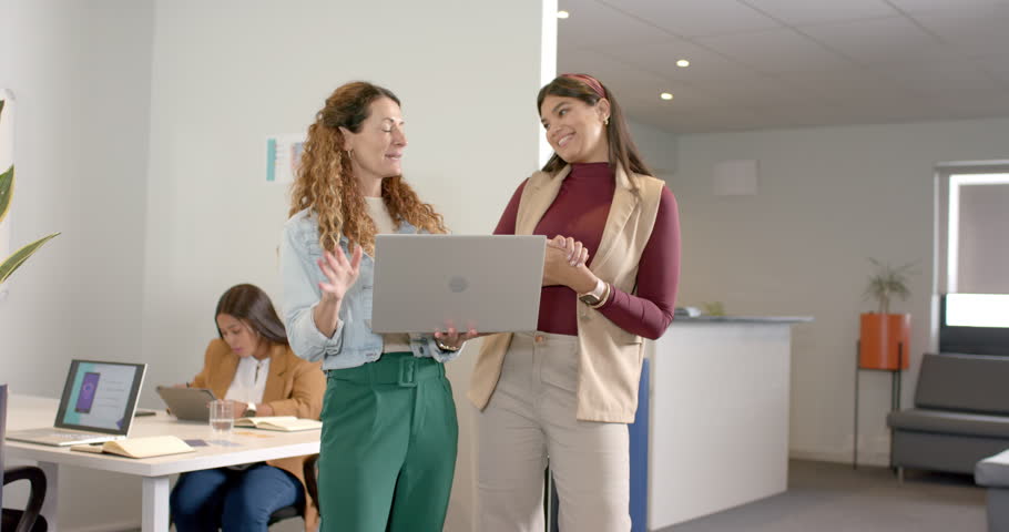 Diverse female coworkers tapping laptop trackpad and guiding project demo in open-plan office. Collaboration, professionalism, teamwork, innovation, productivity, modern, dynamic