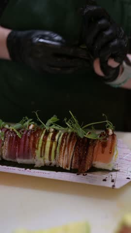 Chef garnishing colorful sushi roll with microgreens and sauce in a professional kitchen, featuring tuna, avocado, eel, and salmon toppings