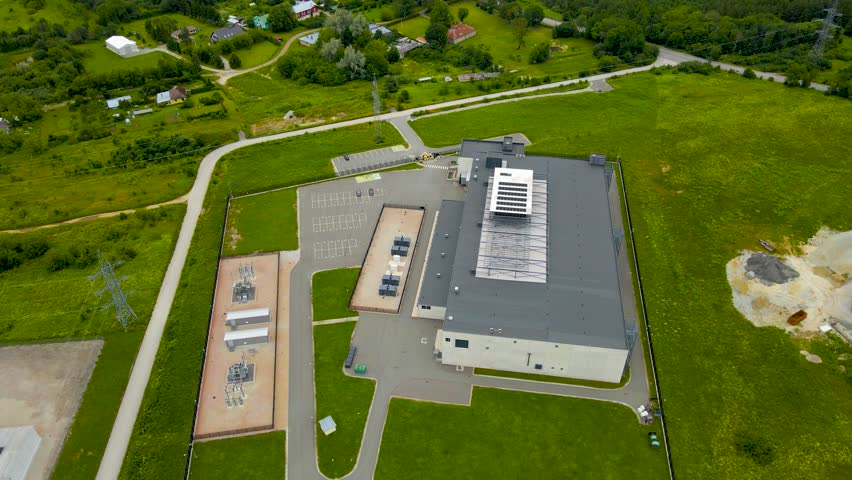 Aerial drone footage orbiting around a gray colored server room or data centre in green grassy countryside that is fenced and has roads around it. Cooling system on the rooftop to cool the server room
