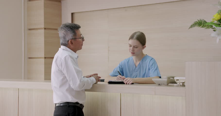 Asian male standing at hospital front desk preparing to pay with credit card while female nurse recording payment details during healthcare billing process