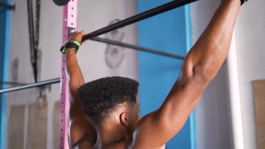 Close-up of muscular Black man's sweaty back and defined arm muscles during intense pull-up exercise with green wrist straps in modern functional training gym