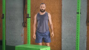 Athletic bearded man performing dynamic box jump exercise in functional training gym with gray tank top and blue shorts surrounded by green squat rack  - Powered by Shutterstock - Get 15% off with code: PIKWIZARD15