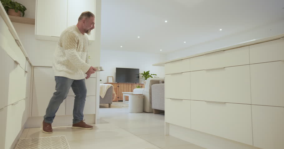 Carrying metal bowl mature man kneeling by cabinet guiding puppy onto mat feeding in bright kitchen. Domestic, caregiving, affectionate, homey, cozy, family, lifestyle