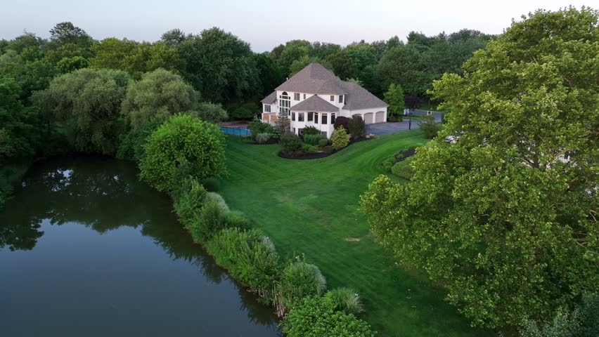 Aerial view of a large upscale American home with multi-gabled roof, expansive windows, landscaped garden and backyard pool, set in peaceful suburban neighborhood. Approaching shot.