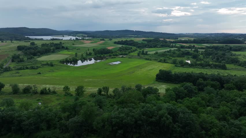 Green farmland in American during cloudy day in summer. Aerial wide shot. Lake and farmstead in background. Green grass and meadows.