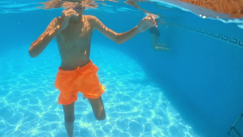 Young boy diving, wearing orange shorts and mask, making playful underwater gestures in bright swimming pool
