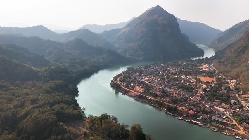 Aerial view of Nong Khiaw, Laos, where the Nam Ou River curves past a small riverside town nestled among lush green mountains and dramatic limestone peaks under a hazy sky