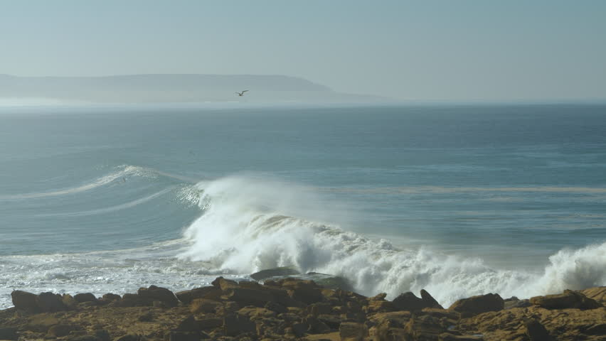 SLOW MOTION: Large cresting wave crashes against the rocks, raising a huge cloud of spray. Hazy horizon and distant coastline further emphasize the sense of the size and vastness of the wavy ocean.
