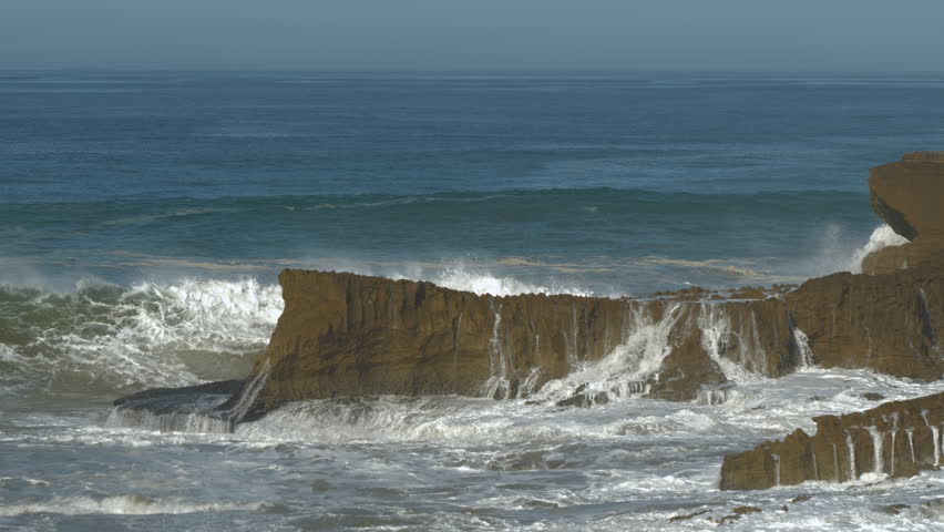 SLOW MOTION: Huge wave crashes against a cliffside, sending enormous amount of white water and foam high into the air. Water cascades down the face of a scenic cliff and spills over the rocks below.