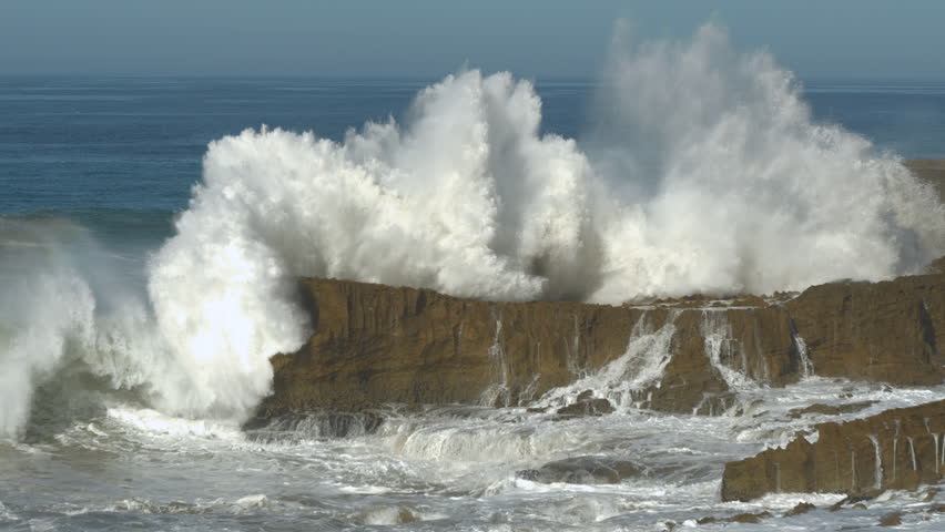 SLOW MOTION: Huge wave crashes against a cliffside, sending enormous amount of white water and foam high into the air. Water cascades down the face of a scenic cliff and spills over the rocks below.