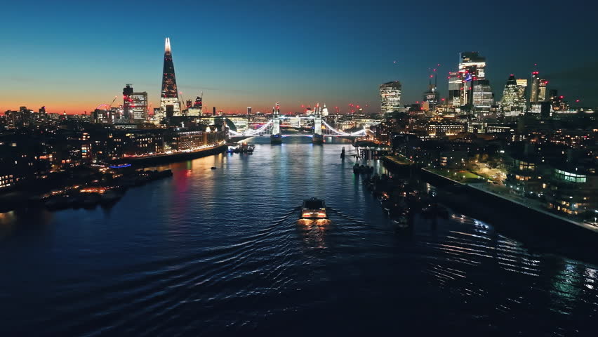 Illuminated London city skyline at sunset, Tower Bridge, The Shard, and a ferry navigating the River Thames. Night lights cityscape. United Kingdom, Europe. Travel destination. Drone flight footage