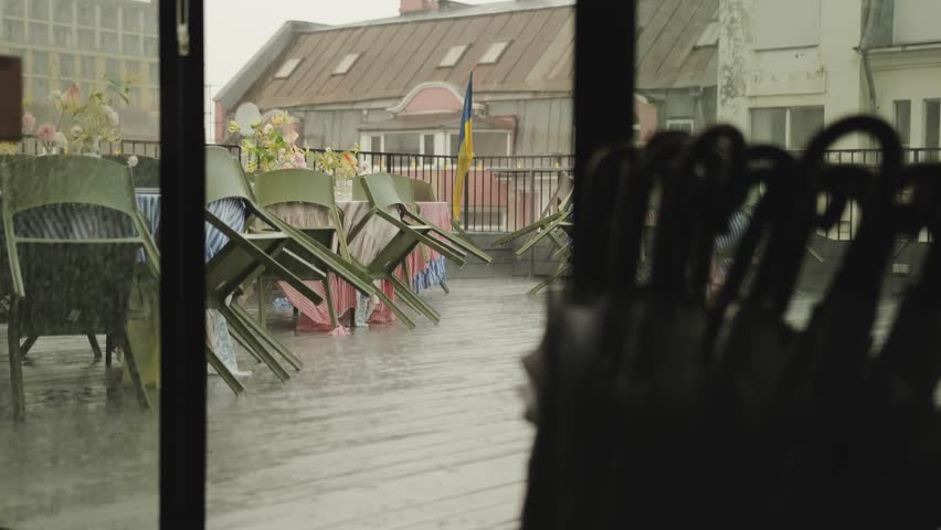 View through sliding doors of a cafe terrace during the rain. Empty wet tables decorated with flowers, with green chairs. The restaurant is closed due to the weather.