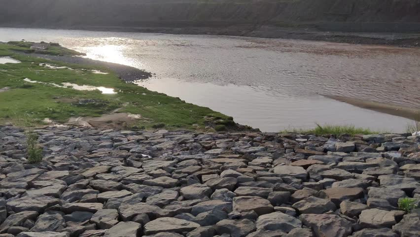 Flowing river under bright sunlight reflecting light beside steadfast mountain stones in peaceful landscape