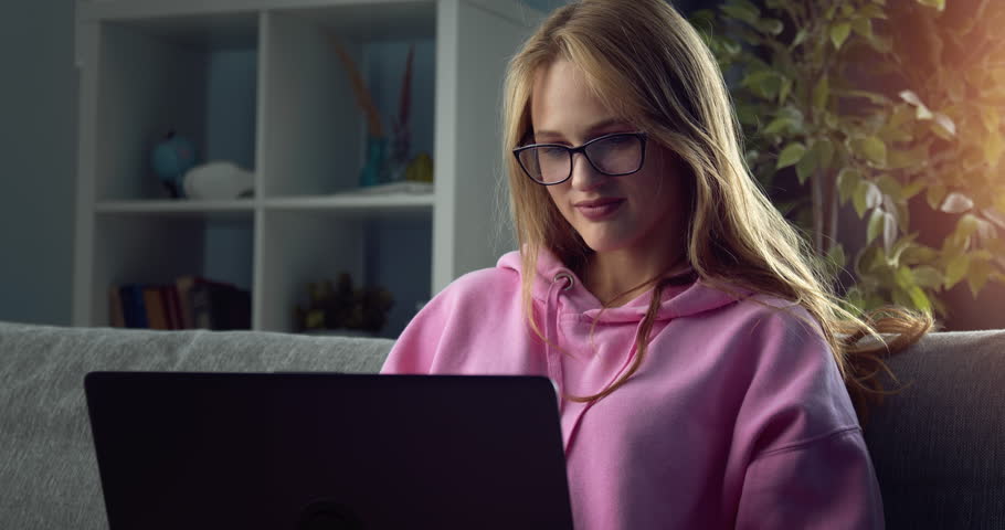 Charming young woman in eyeglasses and pink hoodie having video chat on wirless laptop. Relaxing on comfortable couch at home and having online conversation.