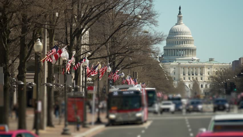 Colorful flags decorate Pennsylvania Avenue, leading to the iconic US Capitol building, as vehicles move through the bustling street on a clear day.