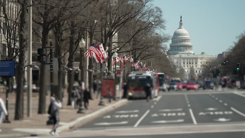 Colorful flags decorate Pennsylvania Avenue, leading to the iconic US Capitol building, as vehicles move through the bustling street on a clear day.