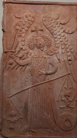 Vertical screen ancient carved stone relief depicting an angel with large wings holding scales and a cross surrounded by symbolic figures representing a scene of judgment or balance