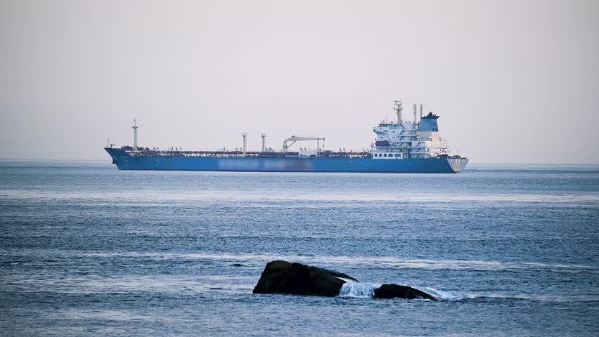 A large blue cargo ship anchored offshore of Keelung Waimushan, Taiwan. Waves crash gently against the rocks, creating a peaceful seaside scene in the morning.