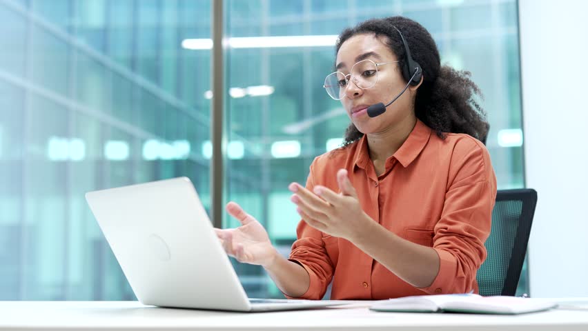 Confident african american businesswoman in wireless headset talking on video call conference using a laptop sitting in office. Female coach speaks remotely, communicates at online meeting, training