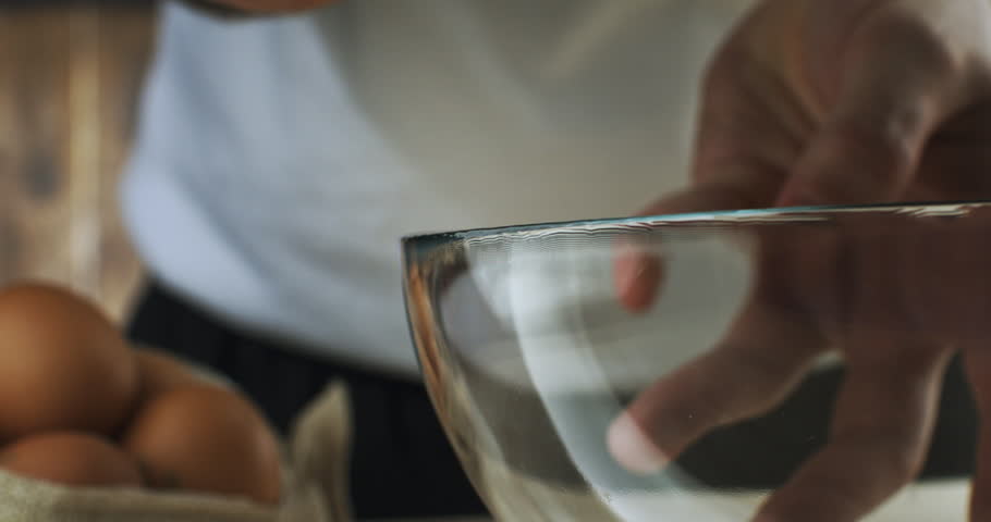 Super Slow Motion Macro of Hands Cracking a Fresh Brown Egg on the Edge of a Transparent Glass Bowl. Real-Time Scene of Cooking or Baking Preparation in a Rustic Kitchen with Natural Ingredients.