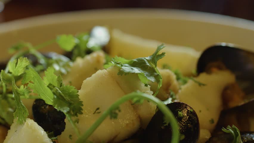 A close-up view of a hand using a spoon to serve hot seafood chowder with mussels, squid, scallops, and fresh cilantro in warm, natural lighting