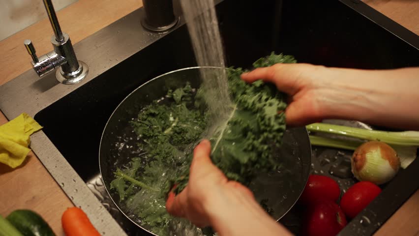 Professional chef rinsing organic kale under stream of water, preparing fresh ingredients for nutritious meal with colorful vegetables scattered on clean kitchen counter