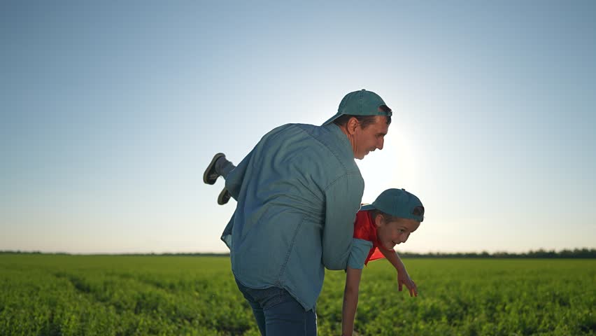 Father carries boy pretending superhero across green field. Happy boy in red cape smiles. Joyful outdoor play with father. Boy enjoys adventure superhero. Child laughs flying with father superhero.