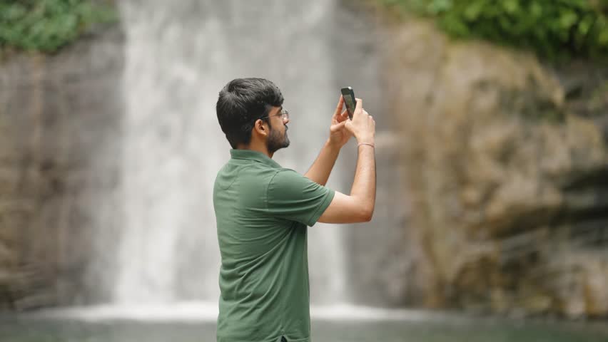Indian man taking photos with smartphone near waterfall in mountains, scenic nature background, 4k video