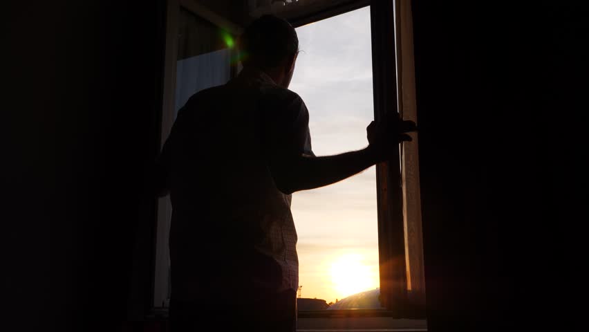 Man unveil curtains and open window, stand and look out to sunset and city. Dark room, silhouetted low angle shot. Bright evening sun shine through open window