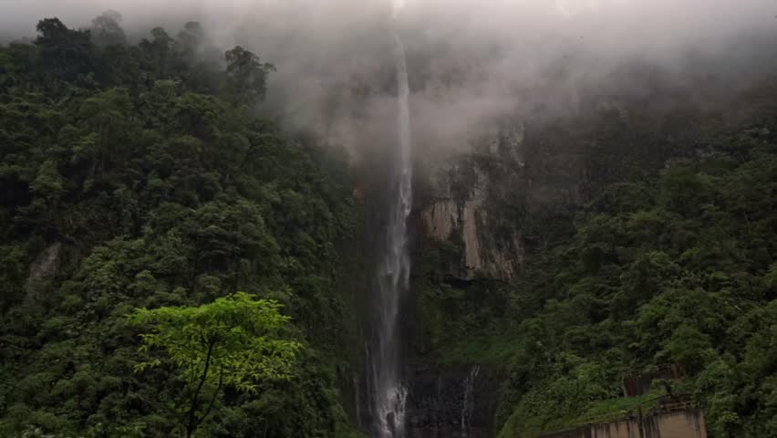 Waterfall cascading down a high, rocky cliff face along the trail to Quebrada Gata. The surrounding lush, green rainforest is partially obscured by fog, creating a mystical and immersive atmosphere.