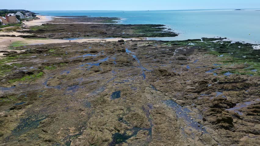 Panning drone movement over the Plage de Bonne-Source tidal shoreline with algae-covered rocks and shallow streams, Pornichet, Loire-Atlantique, France.