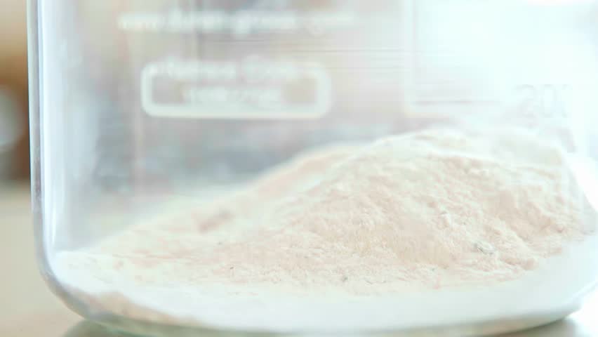 A macro close-up shot shows a white chemical powder being added to a glass beaker in a clean, modern laboratory for a scientific or pharmaceutical experiment.