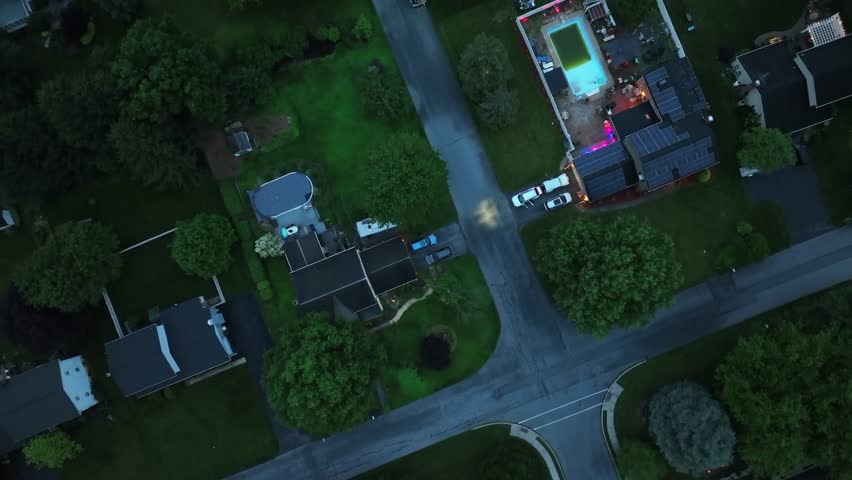 Aerial night view of American suburban street with glowing backyard lights, lit-up swimming pool, and surrounding houses. Peaceful summer evening in quiet residential neighborhood. Top down.