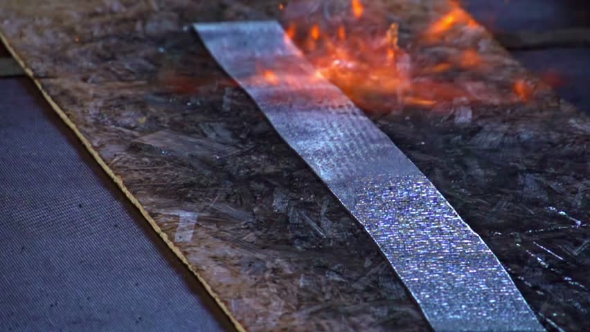 An industrial blowtorch applies intense heat and an open flame to a fireproof material strip to test its durability, heat resistance, and fire-retardant properties in a close-up demonstration.