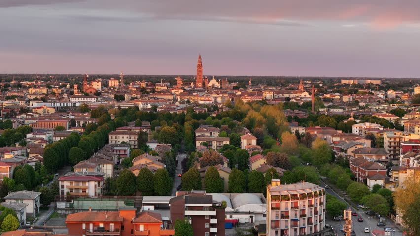 Aerial slow drone pull-away reveals the iconic skyline of Cremona, with the cathedral and medieval towers glowing in golden evening light above tree-lined avenues and city rooftops.