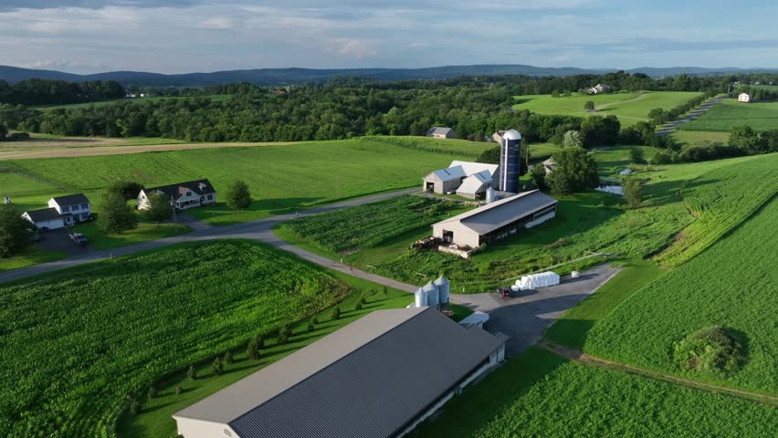 Aerial view of American farm in summer. Large barns, silos and lush green fields stretch across rolling hills. Farmhouses and rural roads complete peaceful countryside landscape. Descend shot.