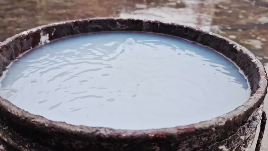 Rainwater falls into a blue bucket, creating splashes and ripples. Slow motion captures raindrops and objects impacting the water surface, emphasizing texture and movement