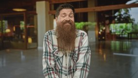 Bearded man in green plaid shirt smiling and leaning forward inside hotel building lobby near reception desk; friendly welcome warmth hospitality. - Powered by Shutterstock - Get 15% off with code: PIKWIZARD15
