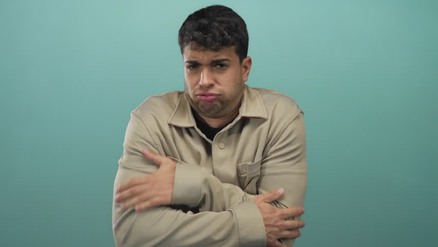 Man firmly hugs arms against background while hispanic young male poses in portrait wearing shirt teal showing cold discomfort.
