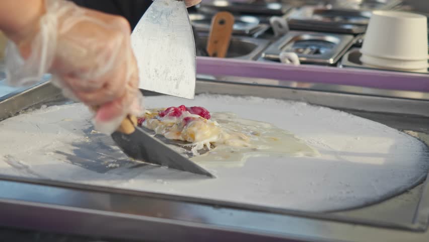 Two hands of skilled street chef using spatulas to mix frozen fried ice cream ingredients of strawberry, cream, and banana on metal plate.