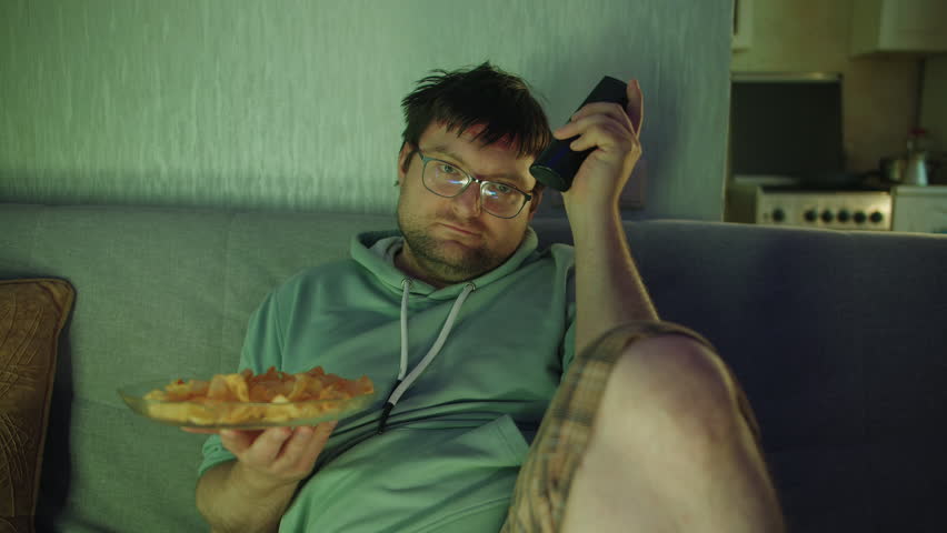 Close up front view of a tired man in home clothes sitting on a sofa holding a plate of chips and changing the channel with a TV remote in a living room at evening. Watching movies as a hobby
