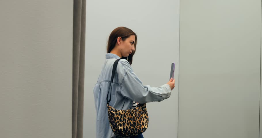 Fashionable customer tries different poses while taking selfies with smartphone in a clothing store's dressing room, using the mirror for a wider shot. concept of shopaholism in women