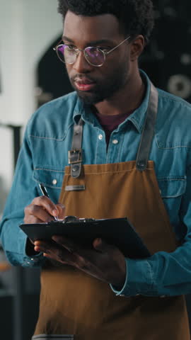 Small business owner African American male guy man working in roasted fabric control roasting coffee beans machine process technician checkup machinery equipment checking quality writes notes data
