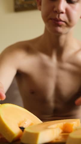 Vertical video of a skinny hungry child eating a ripe juicy melon, greedily biting the sweet pulp of a mature fruit treat. Teen holds melon slice with both hands and eats with good appetite.