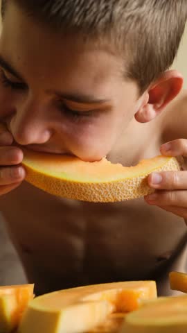 Vertical video of a skinny hungry child eating a ripe juicy melon, greedily biting the sweet pulp of a mature fruit treat. Teen holds melon slice with both hands and eats with good appetite.