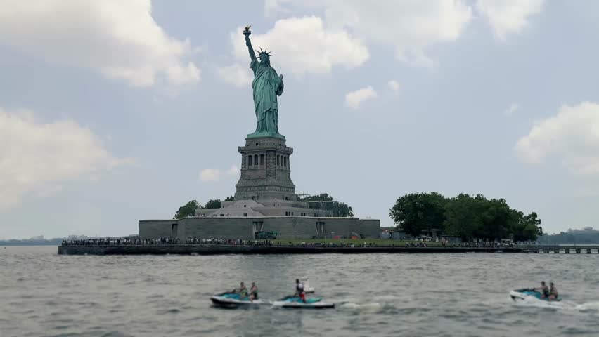 Distant view of the Statue of Liberty on Liberty Island in New York Harbor, with groups of people riding jet skis across the Hudson River in the foreground. Tourists line the perimeter of the island.