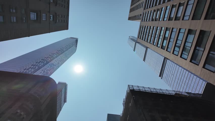 Upward view of Manhattan skyscrapers under a bright midday sun. Modern glass and steel towers contrast with older brick architecture, with a lens flare adding a dynamic visual element against the sky.