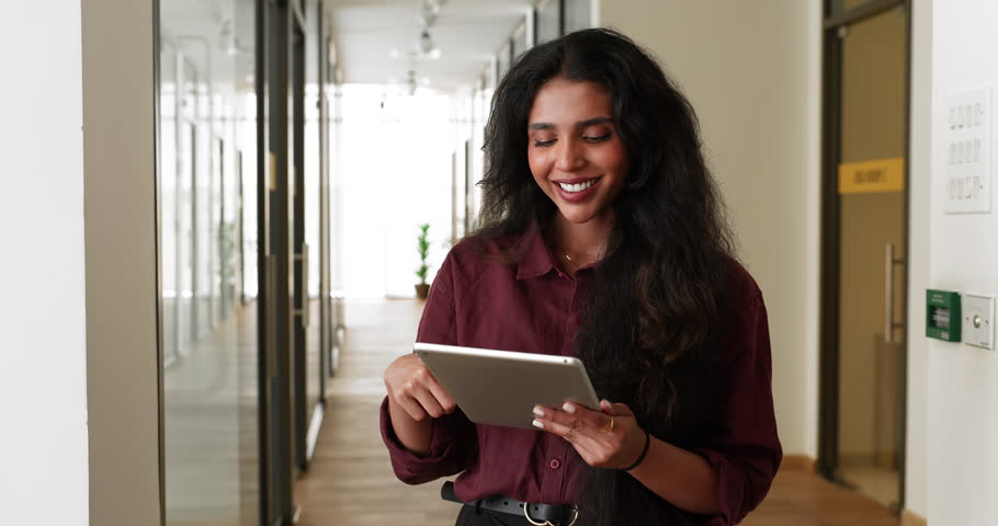 Attractive Indian woman office employee standing in modern workplace hallway using digital tablet, reviewing files, online presentation slides of received document, watching marketing campaign content